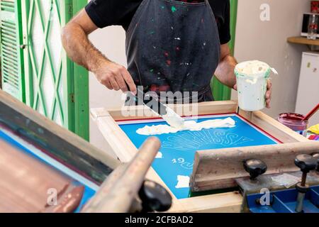 Crop uomo in grembiule sporco lavorando con serigrafia durante la creazione stampa su t-shirt in officina Foto Stock