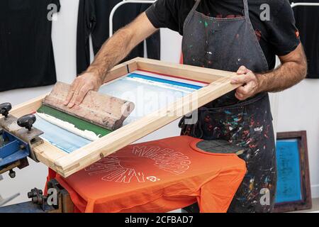 Crop uomo in grembiule sporco lavorando con serigrafia durante la creazione stampa su t-shirt in officina Foto Stock