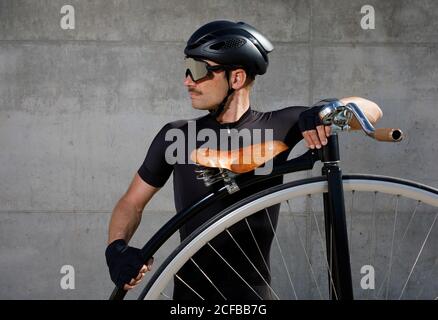 Uomo calmo determinato in abbigliamento nero e casco guardando via in piedi su strada asfaltata e in posizione inclinata sulla ruota alta bicicletta contro muro di cemento in giornata di sole Foto Stock