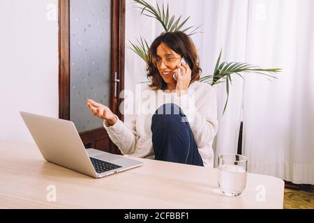 Focalizzato giovane freelance in abiti casual parlando su smartphone mentre si siede al tavolo con il computer portatile e si lavora al progetto a casa Foto Stock