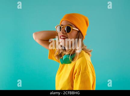 Vista laterale di felice giovane cute femmina in vestito giallo e arancio beanie guardando via indossare occhiali da sole occhiali da sole contro colorati sfondo turchese Foto Stock