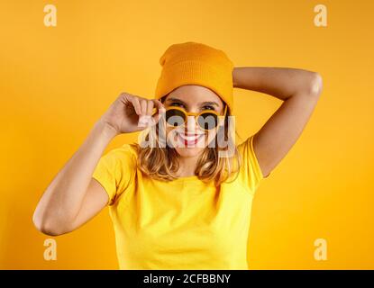 Felice giovane donna in abiti luminosi e occhiali da sole eleganti sorridendo e guardando la fotocamera su sfondo arancione Foto Stock
