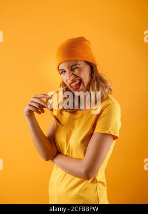 Vista laterale di felice giovane donna in abiti luminosi e tenendo gli occhiali da sole alla bocca sorridendo e guardando la macchina fotografica mentre lampeggiano gli occhi su sfondo arancione Foto Stock