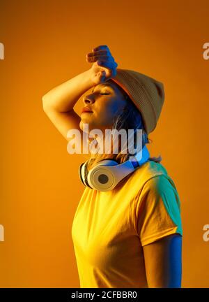Giovane donna premurosa in beanie arancione con cuffie intorno al collo che tengono il braccio sopra la testa con occhi chiusi su sfondo arancione Foto Stock