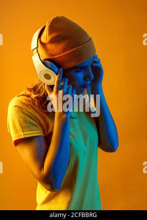 Vista laterale di una giovane donna pensata e sicura in un beanie arancione che ascolta la musica sulle cuffie guardando lontano da sfondo arancione Foto Stock