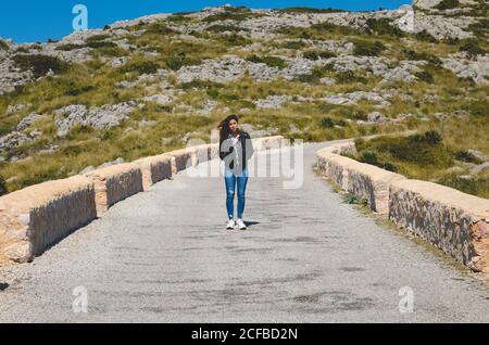 Giovane donna in abito casual tenendo le mani in tasca e. in piedi su un sentiero roccioso vicino alla collina in una giornata di sole natura Foto Stock