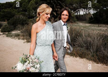 Bella coppia appena sposata in elegante abbigliamento da sposa che tiene le mani mentre camminando sulla natura Foto Stock