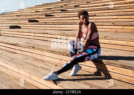 Giovane donna afro-americana slim in colorati abiti sportivi e sneakers bianchi guardando via con curiosità e contemplazione mentre si siede da soli su scale di legno marrone sullo stadio Foto Stock