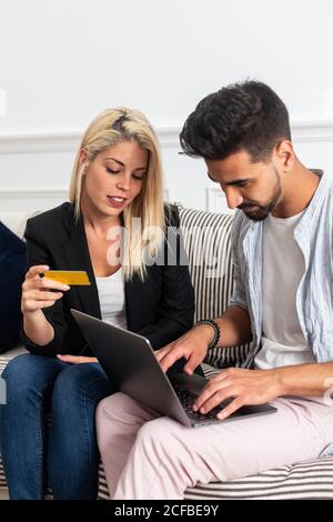 Blonde femmina sorridente e leggere credenziali carta di credito a allegro ragazzo etnico con computer portatile mentre si siede sul divano e fare acquisti online insieme Foto Stock