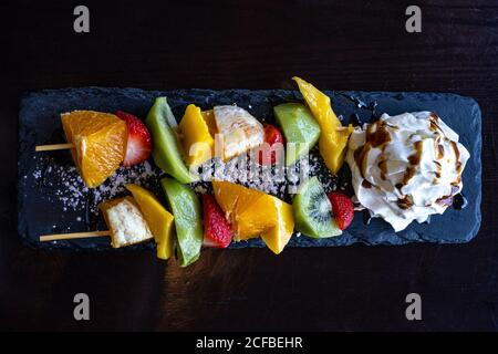 Kebab di frutta fatto di arance, fragole, Kiwi, Mango servito con panna montata. Primo piano della macro. Messa a fuoco selettiva Foto Stock