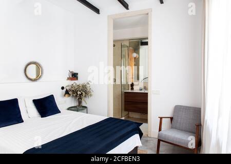 Comodo letto con piumone bianco e blu e morbida poltrona situato vicino alla porta del bagno in una camera da letto accogliente di appartamento moderno Foto Stock