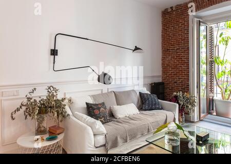 Comodo divano con morbidi cuscini sotto la moderna lampada in metallo vicino a tavoli con decorazioni in elegante e accogliente soggiorno a. casa Foto Stock