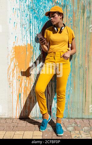 Elegante donna millennial sicuro in elegante vestito giallo brillante e. con le cuffie wireless e lo smartphone mentre ci si trova contro una parete grigia con coloratissime gocce di vernice in giornata di sole Foto Stock