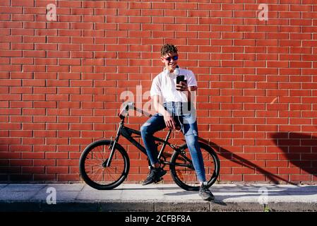 Giovane sorridente afroamericano bell'uomo con capelli afro dentro abito casual seduto in bicicletta e utilizzando smartphone Foto Stock