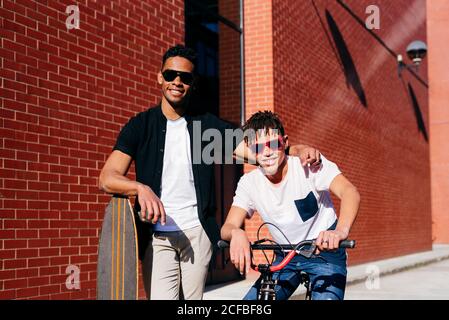 Giovani felici uomini afro-americani bei in abiti casual e. occhiali da sole in piedi sulla strada con bicicletta e skateboard Foto Stock