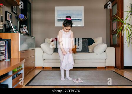 Carino bambina in rosa chiaro leotard e collant mettere sulla gonna mentre ci si trova vicino alle scarpe da ballo in un ambiente accogliente camera a casa Foto Stock