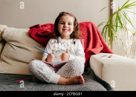 Incantevole piccola ragazza seduta e sorridente sul lettino con un notebook Foto Stock