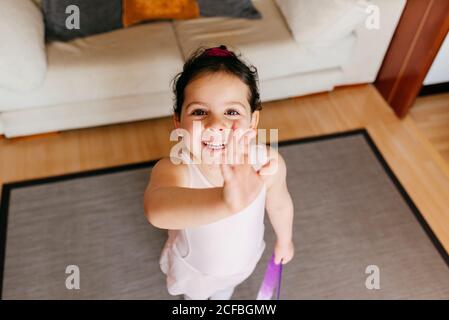 Da sopra bambina con nastro sorridente per macchina fotografica e. agitando la mano durante la ginnastica ritmica a casa Foto Stock