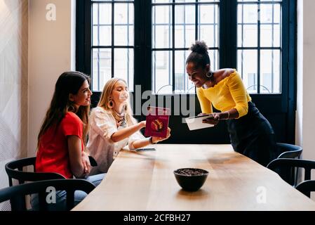 Abbastanza sorridente giovane cameriera afroamericana che prende ordine dal moderno le amiche si siedono al tavolo accanto alla finestra del caffè e puntando al menu Foto Stock