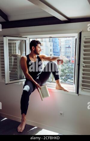 Vista laterale di un giovane uomo bello e serio in casual senza maniche camicia seduta sulla finestra e lettura interessante libro Foto Stock