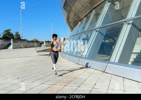 Giovane snella donna afroamericana attiva in abbigliamento sportivo che corre lungo costruzione della città durante l'allenamento nella soleggiata giornata estiva Foto Stock