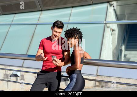 Personal trainer che usa lo smartphone e mostra l'app al fitness sorridente Donna afro-americana mentre si sta insieme accanto alla costruzione in giorno estivo soleggiato Foto Stock