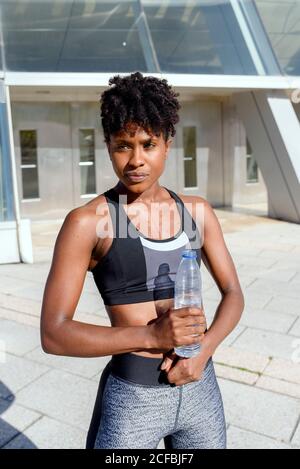 Giovane allegra donna froamericana corridore in top sport e. leggings in piedi accanto alla costruzione e acqua potabile mentre hanno pausa durante l'allenamento in città Foto Stock