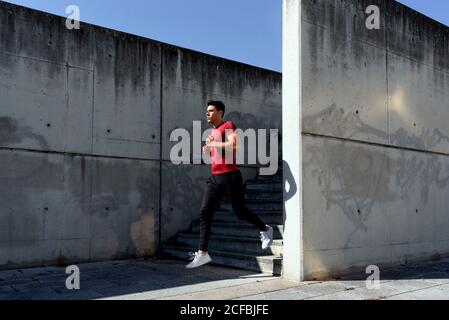 Forte sano maschio attivo in camicia rossa e pantaloni neri scendendo le scale durante l'allenamento nella soleggiata giornata estiva città con muro di cemento sullo sfondo Foto Stock