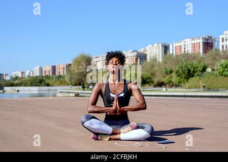 Moderno sportivo afro-americano seduto in posizione lotus e meditating dopo l'allenamento in riva al fiume in città in estate giorno di sole Foto Stock