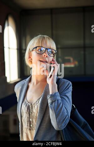 Giovane donna d'affari bionda in occhiali che parla sullo smartphone e guarda via Foto Stock