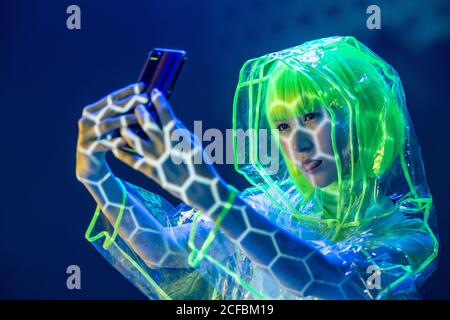 Giovane donna asiatica in abbigliamento futuristico e parrucca verde prendendo selfie su smartphone in luce fluorescente Foto Stock