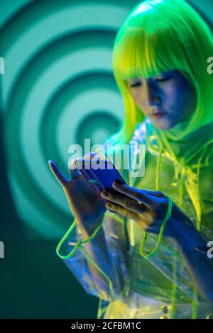 Giovane donna asiatica in abbigliamento futuristico e parrucca verde utilizzando smartphone in luce fluorescente Foto Stock