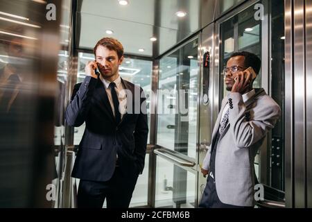 Due multinazionali del business man standing in ascensore e utilizza lo smartphone. La gente di affari in grande ascensore di vetro in un ufficio moderno Foto Stock