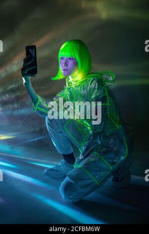 Giovane donna asiatica in abbigliamento futuristico e parrucca verde prendendo selfie su smartphone in luce fluorescente Foto Stock