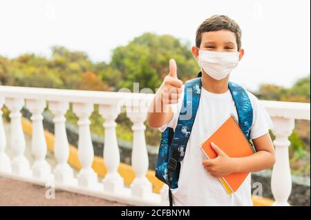 Ritratto di ragazzo di scuola indossando maschera protettiva per l'inquinamento o virus e mostrando pollici in su mentre in attesa di bus di scuola al mattino. Coron Foto Stock