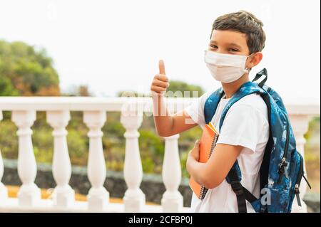 Ritratto di di bambino di scuola che indossa maschera protettiva per l'inquinamento o virus e mostrando pollici in su. Coronavirus e concetto scolastico Foto Stock