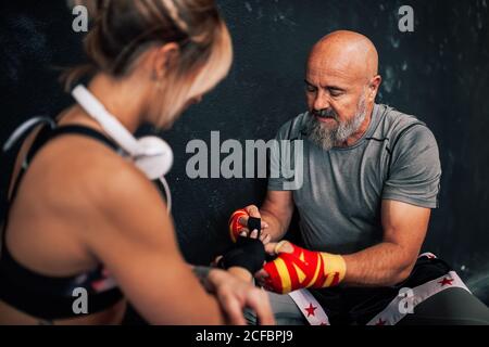 Istruttore maschile bearded adulto che aiuta l'atleta donna a fasciare al polso mentre si siede vicino alla parete nera e si prepara per l'esercizio in palestra di pugilato Foto Stock