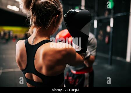 Vista posteriore dell'atleta sportivo giovane e irriconoscibile in guanti da boxe allenarsi combattendo con anonimo allenatore maschile professionista in palestra moderna Foto Stock