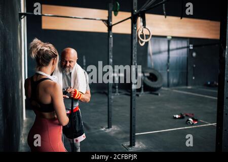 Adulto bearded maschio istruttore aiutare l'atleta donna che avvolge il bendaggio sul polso mentre si sta preparando per l'esercizio in palestra di pugilato Foto Stock