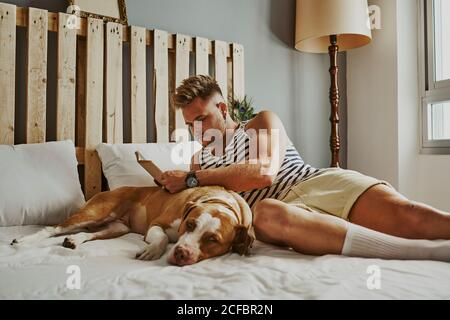 Un ragazzo biondo giovane che legge a letto con il suo cane. Stile di vita concetto Foto Stock