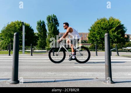 Vista laterale del maschio atletico in occhiali da sole con camicia bianca e pantaloncini neri in bicicletta sulla strada della città con verde alberi sulla strada il giorno d'estate con cielo blu Foto Stock