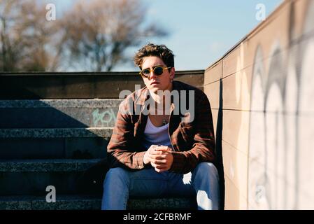 Giovane bel ragazzo in abiti casual guardando la macchina fotografica mentre seduto sulle scale nella giornata di sole sulla strada della città Foto Stock