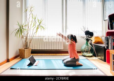 Vista laterale della bambina positiva seduta sulle ginocchia in yoga posa sul tappetino vicino alla finestra a casa mentre praticare yoga e guardare video tutorial sul tablet Foto Stock