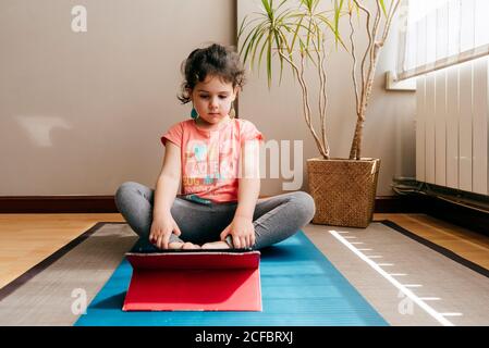 La bambina premurosa si siede sul tappetino vicino alla finestra di casa riposarsi dopo aver praticato lo yoga durante le esercitazioni video sul tablet Foto Stock