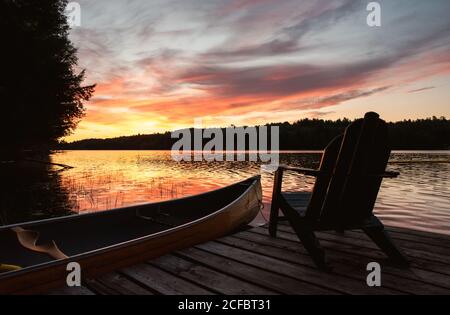 Canoa e sedia adirondack su un molo su un lago all'alba. Foto Stock