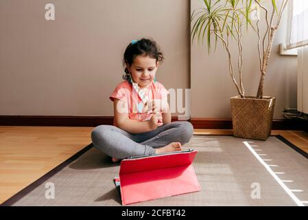 La bambina premurosa si siede sul tappetino vicino alla finestra di casa riposarsi dopo aver praticato lo yoga durante le esercitazioni video sul tablet Foto Stock