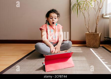 Sorprende la bambina che si siede sul tappetino vicino alla finestra di casa riposarsi dopo aver praticato lo yoga durante le esercitazioni video sul tablet Foto Stock