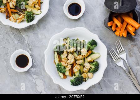 Da sopra di insalata fatta di pollo fritto e. zucchine con carote e broccoli e poste in ciotola vicino salsa di soia e forchette sul piano di marmo Foto Stock