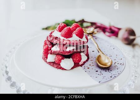 Angolo di deliziose frittelle di barbabietola biologiche con lampone fresco su piatto con cucchiaio dorato su tavola bianca Foto Stock