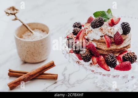 Gustosi pancake con fragole fresche e more decorate con menta foglia posta sul piatto vicino a bastoni di cannella e tazza di gelato su tavolo di marmo Foto Stock